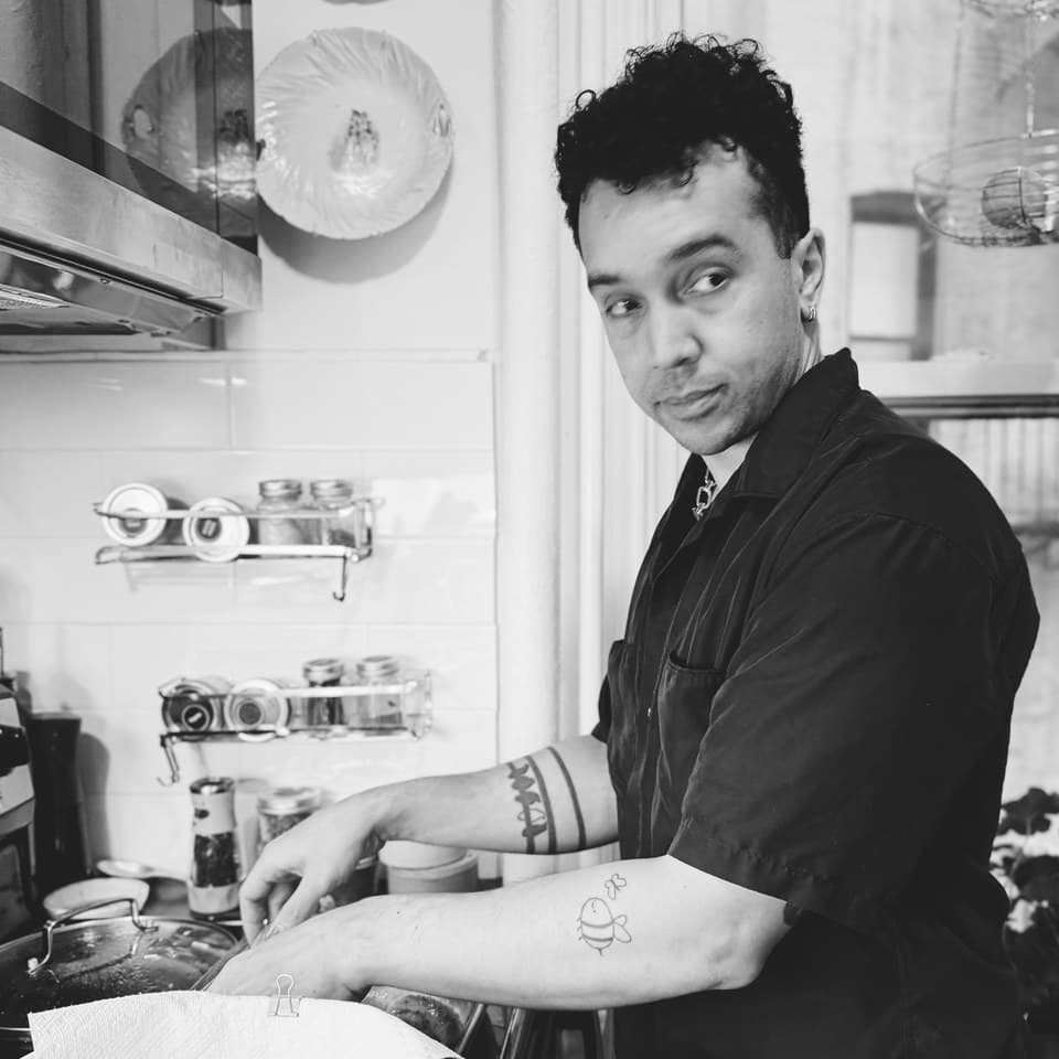 Ry in a kitchen, turning from the stove toward the camera — black shirt, forearm tattoos, shelves of jars and pans behind him
