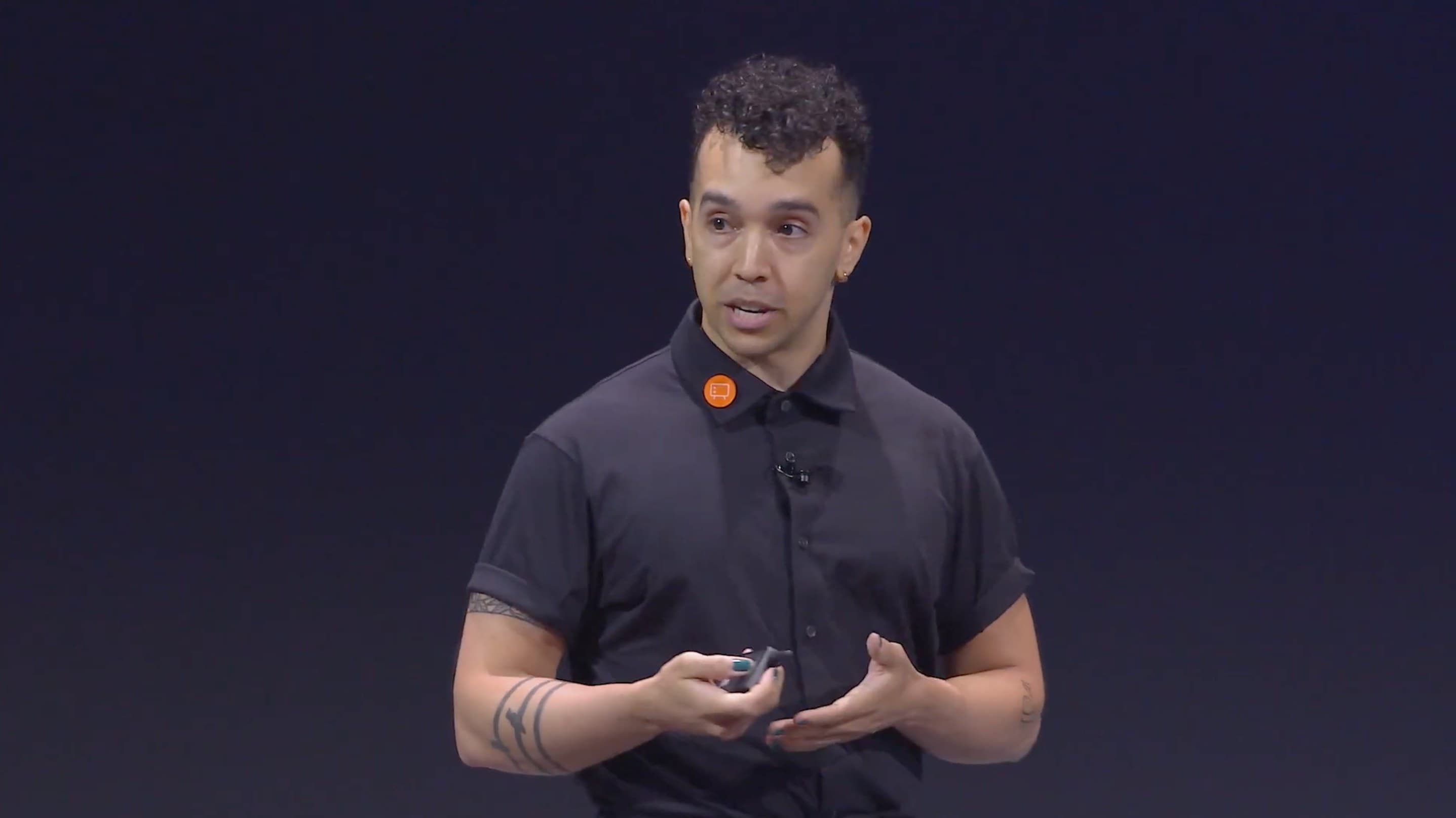 Ry mid-sentence on a darkened conference stage, in a black polo with a clicker in hand and a small orange Config pin on the collar